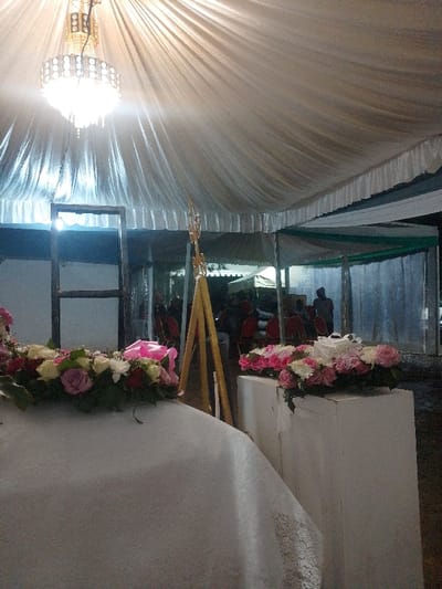 The image captures an event space in Marenyo-Shianda ward, Kenya, illuminated by artificial light. A large, ornate, multi-tiered chandelier hangs centrally from a ceiling draped with white, pleated fabric. In the immediate foreground on the left, a white-draped surface displays a linear arrangement of pink, white, red, and purple roses with green foliage, featuring a pink ribbon. Adjacent to this, a golden, multi-pronged decorative stand with a tripod base is positioned. To the right, a white rectangular pedestal supports another floral arrangement of pink and white roses, within which a small white gift box with a white ribbon is placed. Beyond these elements, the background reveals dark-framed structures. Indistinct human figures are visible in the mid-to-far background, some seated in what appear to be red chairs, and others standing. No specific actions or interactions among the individuals are clearly discernible. The overall environment suggests a prepared setting for a gathering or celebration.