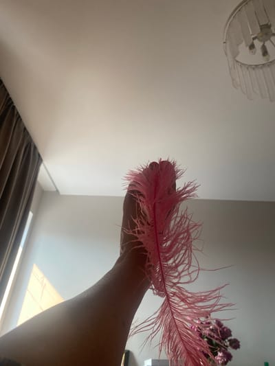 A hand holds a large, pink feather, its fluffy barbs fanned out. The background is a soft white ceiling, with a light fixture visible in the upper right corner. To the left, a dark brown curtain is drawn. In the lower right, a blurred bouquet of purple flowers is visible. The scene is indoors, likely a room in Novi Petrivtsi, Ukraine. The lighting appears to be natural, coming from a window not visible in the frame, casting soft shadows.