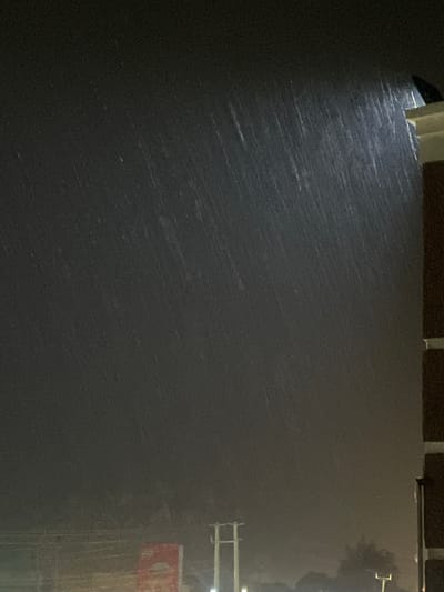 The image depicts a night scene during heavy rain. The location is likely Hayin Banki, Nigeria, as specified. Rain streaks are visible across the frame, illuminated by a light source in the upper left. A building corner is visible in the upper center and left of the image. A streetlight and a sign are present on the right. The overall environment suggests an urban or suburban setting, with limited visibility due to the rain and darkness.