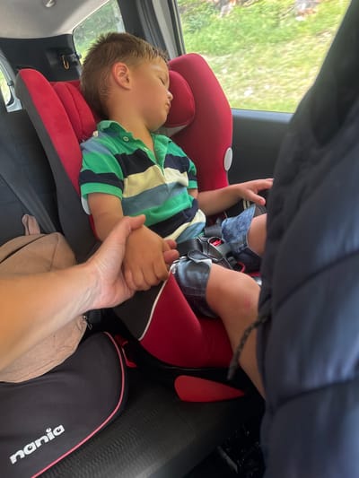 A young boy, asleep in a red car seat, is in the back of a vehicle. He is wearing a striped polo shirt and denim shorts. His right hand is clasped by an adult's hand. A light brown bag is visible to the left of the car seat, and a black bag with the brand "nania" is on the floor. The background outside the car window shows green foliage, suggesting a rural or suburban setting, possibly near Longchaumois, France. The overall scene depicts a peaceful moment of a child resting during a car journey.