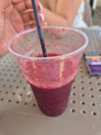 A close-up, eye-level shot shows a clear plastic cup filled with a dark purple berry smoothie, with red berry seeds visible on the inner surface of the cup. A black plastic straw is partially submerged in the smoothie. A hand with rings is visible on the left, holding the cup. In the background, out of focus, a person in a white top and a pack of cigarettes are visible. The scene appears to be outdoors on a table with a speckled texture. The overall context suggests a casual setting, possibly at a cafe or an outdoor seating area in Ta' Xbiex, Malta.