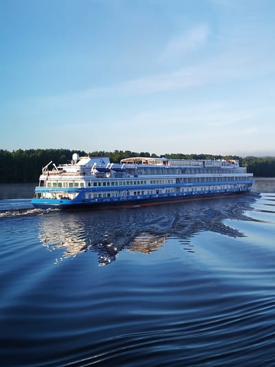 The image depicts a large, multi-deck river cruise ship, identified by the name "РОССИЯ" (Russia) prominently displayed on its white and blue hull, navigating a calm river. The ship's lower hull is blue with a distinctive orange stripe at the waterline. Multiple decks feature rows of rectangular windows, indicating passenger cabins, and several open deck areas are visible, some with white awnings. Lifeboats are stowed on the upper decks, and communication equipment, including radar domes and antennas, is visible on the superstructure. A flag, likely Russian, is flying from the stern. The ship is moving from left to right, creating a distinct V-shaped wake with turbulent white foam immediately behind it, which then dissipates into smooth ripples. The ship's reflection is clearly visible in the dark blue water, distorted by the surface ripples and mirroring the vessel's structure and colors. The background features a dense, dark green forest lining the riverbanks. A faint, low-lying mist or fog is present above the distant treeline, suggesting an early morning scene. The sky above is a clear, light blue with sparse, wispy white clouds. The scene is set on a river in Vasil'ki, Russia. No individuals are discernible in the image.