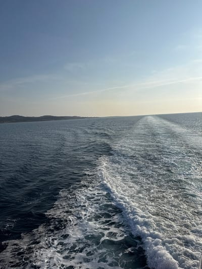 The image displays an open-water scene near Kamariotissa, Greece, viewed from the stern of a moving vessel. The lower two-thirds of the frame are occupied by dark blue-green sea water, featuring a prominent, frothy white wake trailing directly behind the point of view and extending towards the center-right horizon. Ripples and small waves are visible across the water surface. A distant, low-lying coastline, characterized by undulating terrain and dark green vegetation, occupies the left side of the mid-ground. The upper third of the image depicts a clear, light blue sky, which brightens towards the right horizon, indicating the general direction of the sun. Two faint, thin contrails are discernible in the upper sky. No individuals or other specific objects are visible within the frame, beyond the natural environment and the water disturbance created by the unseen vessel.