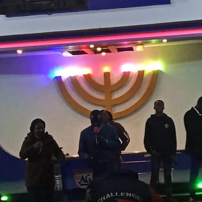 The image depicts an indoor gathering on a stage in Ndeiya, Kenya. A large, golden, seven-branched menorah-like symbol is centrally displayed on a white wall, illuminated by horizontal strips of red, blue, green, and purple LED lights positioned above it. On the stage, a dark-skinned woman in a dark coat and a dark-skinned man in a blue jacket are actively speaking into separate microphones, with the man positioned behind a blue and white podium featuring an "AG" logo. To the right of the speakers, another dark-skinned man in a black hooded sweatshirt with a green star-like emblem stands. Additional individuals are partially visible behind the central speaker and to the far right of the stage. In the immediate foreground, the back of a person's head and shoulder are visible, wearing a dark top with the word "CHALLENGE" partially legible, indicating an audience perspective.