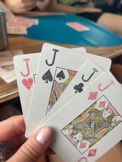 A close-up image taken in Kargat, Russia, depicts a person's left hand holding four playing cards fanned out. The visible cards are the Jack of Hearts, Jack of Spades, Jack of Clubs, and Jack of Diamonds, displayed from left to right. The cards appear worn, with slight discoloration and visible texture. The hand has manicured fingernails with a light pink or nude polish and wears a clear, possibly glass or plastic, ring on the little finger.
The background is out of focus, indicating a shallow depth of field. A light brown wooden surface, likely a table, is visible, with several other playing cards scattered on it to the left. A metallic object, possibly a cup or mug, is partially visible on the far left of the table. In the mid-ground, a blue plastic object, possibly a tray or a container lid, is present, with another card, a red suit, lying on it. Beyond the table, the blurry forms of at least two other individuals are discernible, with arms and partial torsos visible, suggesting multiple participants in a card game. One person wears a light green top and a turquoise bracelet. The overall scene indicates an indoor setting where a card game is in progress.