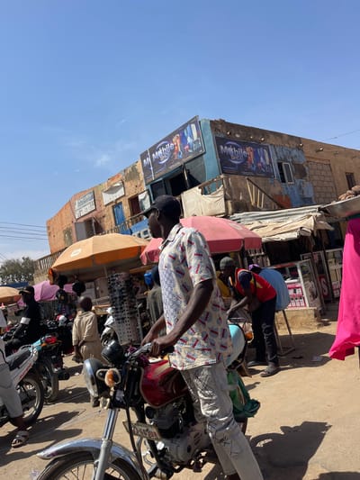 The image depicts a bustling street scene in Chikaji, Nigeria, under a clear blue sky. In the immediate foreground, a dark-skinned male wearing a black baseball cap, a light-colored short-sleeved patterned shirt, and light-colored jeans with dark crosshatch patterns stands straddling a red and silver "BOXER" brand motorcycle. The motorcycle bears a license plate reading "KD-63UN".
Behind this individual, the scene transitions into an active market area. Numerous people are visible, some walking, others engaged at stalls. An orange market umbrella provides shade over a stall displaying multiple rows of sunglasses. Another smaller purple umbrella is visible further back. To the right of the foreground motorcycle, a dark-skinned male wearing a red vest over a blue shirt and dark pants is attending to a motorcycle or stall. On the far right edge of the frame, a person wearing a bright pink garment holds a silver bowl on their head, partially out of view.
In the mid-ground and background, multiple rustic, multi-story buildings with faded paint and visible structural wear define the street. Prominently affixed to one building are two large rectangular signs for "Mobile Shop," featuring images of smartphones and other electronics. Below these signs, makeshift market stalls with corrugated metal roofs and various goods extend along the street. The ground appears to be unpaved and dusty, consistent with a dry, outdoor market environment. Sunlight is bright, casting distinct shadows.