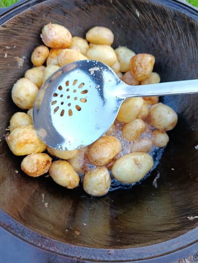 In the village of Zipunovo, Russia, a close-up shot shows a metal slotted spoon scooping small, whole potatoes from a dark wooden bowl. The potatoes appear to be cooked, with some showing golden-brown browning. The bowl is weathered and shows signs of use. The background is blurred, but hints of green suggest an outdoor setting. The lighting is natural, and the image is taken from a high angle.