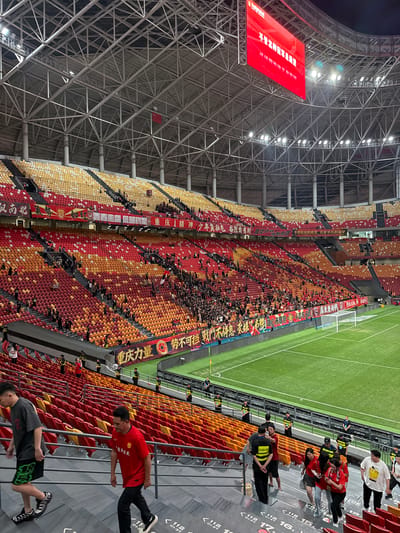 Here is a description of the image:

The image depicts a large stadium in Longxing, China, likely during a sporting event, perhaps a soccer match. The stadium is mostly full, with crowds of people filling the red, yellow, and orange seats. Banners with Chinese characters are displayed along the sides of the seating area. A large red screen with Chinese writing hangs from the stadium's metal framework. A green field is visible to the right, indicating a sports pitch. The lighting suggests it could be evening or a night event. People can be seen walking in the foreground, suggesting movement within the stadium environment. Several individuals, some appearing to be security personnel in uniform, stand near the edge of the seating area.
