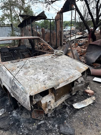 The image depicts a scene of extensive destruction in Nedryhailiv, Ukraine. In the foreground, a severely damaged, burnt-out four-door sedan occupies the center. The vehicle's exterior shows blistered, flaking white and grey paint, with the underlying metal exposed and discolored by heat. The front bumper is distorted, headlights are missing, and the grille area is open, revealing internal components. The hood is intact but heavily scorched. The windshield and all side windows are absent, leaving open frames. The interior is entirely charred, with only the skeletal frames of seats and the steering column visible. A small, light-colored ceramic bowl containing dark, burnt residue rests on the forward section of the hood.

Behind the vehicle, to the left and center, are the ruins of a brick structure, likely a building or garage. Its roof is entirely gone, and sections of the brick walls have collapsed, creating large openings and exposing twisted metal rebar, charred wooden beams, and scattered red bricks. A metal gate or fence structure is partially visible to the left, showing some damage. To the right, charred, bare tree branches frame the upper portion of the scene.

Through an opening in the demolished structure, a white panel van with green lettering is visible in the distant background, parked on what appears to be a street or open area. Further back, green foliage and additional structures are discernible under a partly cloudy sky. The ground immediately surrounding the car and the destroyed building is covered extensively in black ash, charcoal, fragmented debris, and loose red bricks. A single burnt cigarette butt is visible on the ash-covered ground near the front right of the vehicle. There are no individuals present in the scene, which captures the aftermath of a destructive event.