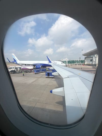 The image, taken from an airplane window, shows an Indigo airline aircraft on the tarmac at an airport, likely in or near Vikram Nagar, India. An Indigo passenger jet, white with blue accents and the Indigo logo, is parked adjacent to a jet bridge. In the foreground, part of the wing of another Indigo aircraft is visible. A fuel truck is positioned next to the parked aircraft, and several baggage carts are also present on the tarmac. A few individuals in high-visibility vests are seen near the truck and carts, indicating ongoing ground operations. The sky above is partly cloudy.
