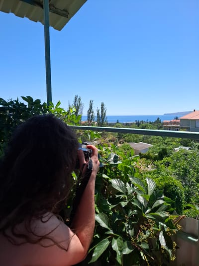A woman with long, dark, curly hair is standing on a balcony and taking a picture with a camera. She is facing away from the viewer and her back is to the camera. The woman is wearing a light-colored top. In front of her, there is lush green foliage, including ivy with small buds. Beyond the foliage and a railing, there is a view of a town with buildings and trees, leading to the blue sea and a clear blue sky. The scene appears to be a sunny day. The location is identified as Щебетовский поселковый совет, Ukraine.