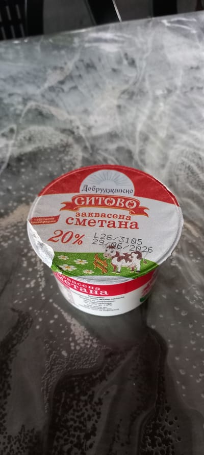 The image shows a container of "Добруджанско СИТОВО заквасена сметана" (Dobrudzhansko Sitovo cultured sour cream) with 20% fat content. The container has a red and white label with a depiction of a cow in a field with flowers and a fence. A date stamp is visible on the label, reading "L26 / 3105 / 29.06 / 2026". The container is placed on a reflective, patterned surface. There is no visible indication of the city of Iskra, Bulgaria, in the image.