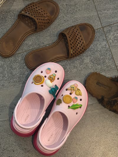 The image shows four pairs of shoes on a gray tile floor. Two pairs are brown woven-style sandals arranged side-by-side, one is a pair of brown fluffy slippers, and the other is two pink Crocs adorned with Jibbitz charms, including a dinosaur, cookie, and cupcake. The photo was taken in a space that is reminiscent of Kerobokan, Indonesia based on the type of flooring and surrounding details.
