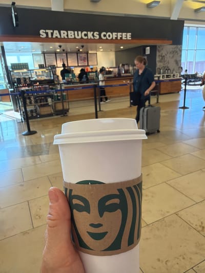A close-up shot shows a hand holding a white Starbucks coffee cup with a brown sleeve featuring the Starbucks siren logo. In the background, a Starbucks coffee shop is visible, with people ordering and staff behind the counter. The location appears to be an airport terminal in Santa Ana, California, USA, indicated by the architecture and the presence of luggage. The environment is bright and modern, with tiled floors and large windows.
