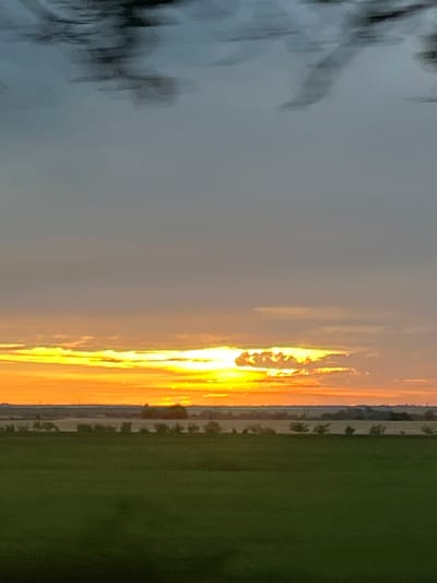 The image depicts a rural landscape at dusk, with a vibrant sunset dominating the sky. The sun's rays create a bright, orange and yellow glow that illuminates a thin strip of clouds near the horizon. Above this, the sky transitions to a pale grey, and further up, dark, blurry clouds streak across the frame, suggesting movement or a distant storm.
The horizon is marked by a line of trees and what appears to be a small structure, possibly a building or a silo, nestled amongst the foliage. The land below is a patchwork of green fields, some of which are covered in vegetation. The overall impression is one of a serene, natural setting, bathed in the warm light of the setting sun.
This image does not contain any information that would suggest it was taken in the city of Slobozia, Moldova, or any other specific geographical location. The scene is generic and could be found in many rural areas.