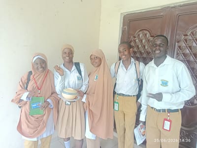 A group of five young people, likely students, stand in front of a brown double door. Two individuals on the left are dressed in light-colored shirts, beige skirts, and headscarves. One of them is holding a green notebook and a beige circular container. The other four individuals are wearing light-colored shirts and beige pants. One person in the middle is wearing a beige headscarf. The two individuals on the right are wearing shirts with a crest on the left side of the chest. One of them is making a peace sign with their fingers. The scene appears to be outdoors, possibly in front of a building. The lighting suggests it is daytime. The timestamp at the bottom right corner indicates the date as 25/06/2026 and the time as 14:36. The individuals appear to be smiling and relaxed, suggesting a positive atmosphere. The location is identified as Unguwar Shanu, Nigeria.