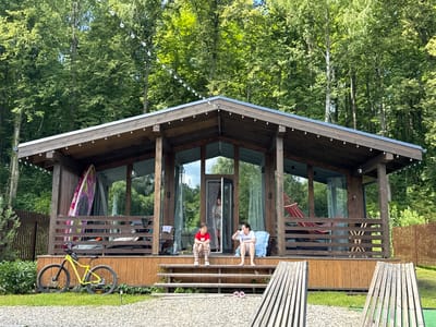 A wide shot captures a modern wooden house with large windows and a porch, nestled amongst dense green trees. The house has a low-pitched roof adorned with string lights. On the porch, a girl in a red shirt and patterned pants sits on the steps, while a woman in a white t-shirt and shorts sits on the porch, drinking from a cup. A surfboard leans against the porch railing, and a yellow bicycle is parked on the grass in front of the house. A red hammock is visible on the right side of the porch, and a wooden fence encloses the property. The scene appears to be a serene, outdoor setting, possibly a resort or private residence in Serpukhov, Russia, judging by the style of the house and the surrounding natural environment.