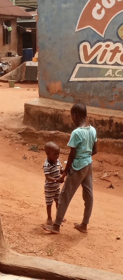 The scene is an outdoor, unpaved area in Umuoji, Nigeria, characterized by reddish-brown dirt. Two children of African descent are present. The child on the right, appearing older, wears a light blue short-sleeved shirt and grey pants, standing with their back mostly to the camera. This child is barefoot. The younger child on the left wears a horizontal striped short-sleeved shirt and matching shorts, facing the older child. This younger child is also barefoot and holds a small, light-colored object in its left hand, while its right hand appears to be in contact with the older child's left hand or wrist.
In the background, to the left, is a building with reddish-brown walls and a reddish corrugated metal roof. A blue plastic barrel and various debris are visible near its base. To the right, a weathered light blue wall features partial white and orange signage. Visible text includes "Co" at the top, "Vita" in the middle, and "A,C," below "Vita." This wall rests on a concrete base. The ground features sparse vegetation in places.