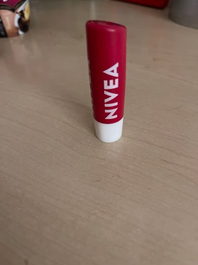 A close-up, eye-level shot shows a red Nivea lip balm tube standing upright on a light brown wooden surface. The tube is positioned slightly off-center to the right. The white base of the lip balm is visible, and the white text "NIVEA" is printed vertically on the red body of the tube. In the blurred background to the left, a portion of what appears to be an ice cream container is visible. The surface is a light wood grain pattern with subtle lines running diagonally from the bottom left to the top right. There are no people or specific Zviahel, Ukraine landmarks identifiable in this image. The lighting is soft, casting minimal shadows.