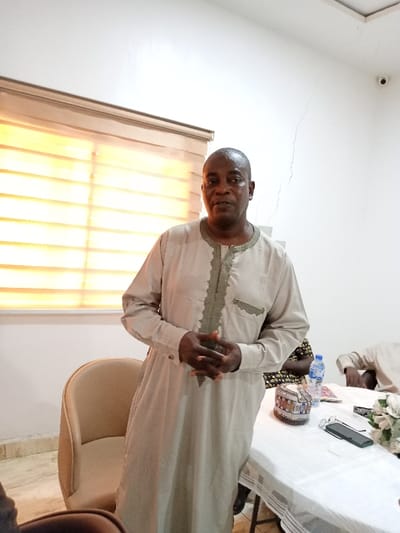 The image shows a man in a long, beige robe with gold trim, standing with hands clasped in front of his chest. He has short, dark hair and a slightly furrowed brow. The man is standing in a room with white walls and a large window with a blind that has been pulled down.  The blinds are light brown in color and are positioned to let in some natural light. The man appears to be speaking to someone off-camera.  The room is sparsely furnished, with a white table in the foreground.  The table is set with a glass of water, a small, round, multi-colored object, and a cell phone.  The coordinates (latitude: 9.198, longitude: 12.491) indicate that this location is in the city of Yola, Adamawa State, Nigeria.  There are no other people in the picture. The man is wearing a simple beige robe and is speaking to another person off camera.  The room is plain and the furniture is simple. There is a white table with a glass of water, a small, round object, and a cell phone on it.  The blinds are letting in a significant amount of sunlight.  A small, round object, likely a hat or purse, is sitting on the table, and a cell phone is partially visible, as are some papers.  There is an object that appears to be a water bottle to the right of the man's shoulder.   The man is standing in a room with white walls, a large window, and a white table in the foreground. The blinds are letting in some light. There is also a brown chair to the left of the man.  The man appears to be speaking to someone off-camera.  The setting is simple and there is no obvious indication of any specific activity taking place.