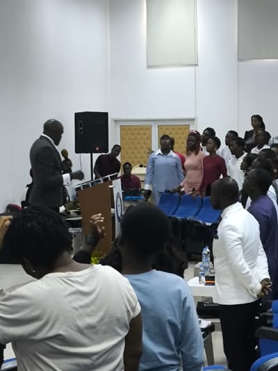 The image depicts an indoor gathering in Kızılbaş, Northern Cyprus / Cyprus, featuring approximately 20-30 individuals within a modern, brightly lit hall with white walls. In the mid-ground, a Black man in a dark suit, white shirt, and tie stands behind a wooden podium displaying a circular logo, holding a microphone in his right hand as if delivering a speech. A large black rectangular speaker on a stand is positioned to his right. To the right of the podium, a group of about ten individuals, predominantly Black and appearing to be young adults, stand facing forward/left, with several looking upward. This group includes a woman in a light blue button-up shirt and glasses, a woman in a peach-colored top, and a woman in a dark red top. Behind this standing group, several rows of unoccupied blue auditorium-style chairs are visible. In the right foreground, a Black man in a white blazer and dark trousers stands with his hands clasped behind his back, facing the direction of the podium. In the immediate foreground, two individuals are seen from behind: a person with dreadlocks in a light-colored top and another in a light blue long-sleeved shirt. A small white table holding three plastic water bottles is present in the lower right. The background features a textured tan-padded wall section, possibly a door, and other individuals, including a man in a dark red shirt near electronic equipment. The environment suggests a contemporary event space or auditorium.