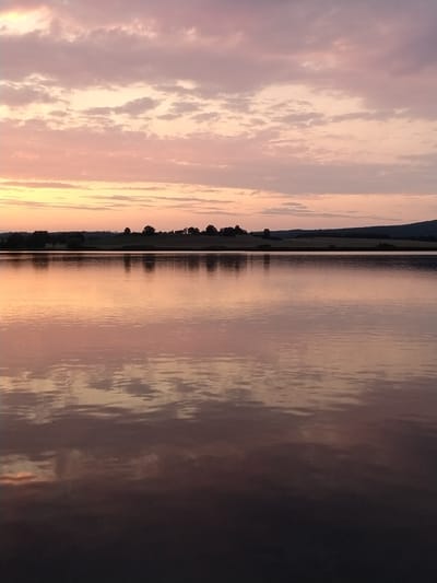 This image shows a serene sunset over a body of water, likely a lake or pond, in Kestřany, Czechia. The sky is a canvas of soft pinks and purples, with scattered clouds catching the warm hues of the setting sun. The water below perfectly mirrors the sky, creating a symmetrical and tranquil scene. The shoreline is visible in the distance, characterized by rolling hills and a line of trees silhouetted against the horizon. The overall atmosphere is peaceful and reflective, capturing the beauty of nature at dusk.