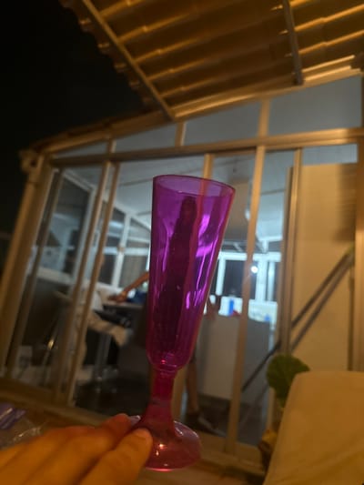 In Palma, Spain, an individual's hand holds the base of a translucent pink stemmed glass. The glass is tall and slender, with a flared rim, and appears to contain a dark, elongated object. The background is a brightly lit indoor space with large windows, revealing what looks like a living area or balcony. The ceiling above features a slatted wooden structure with metallic supports, characteristic of an outdoor covered patio. The lighting suggests it is evening or nighttime.