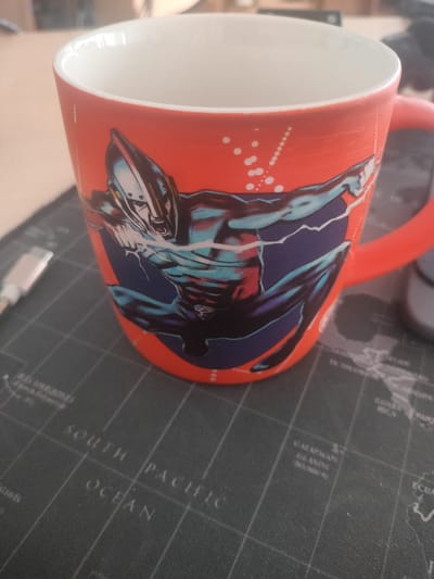 A red mug with a blue and silver superhero design sits on a dark grey mat that appears to be a world map. The superhero on the mug is depicted in a dynamic pose with lightning effects. A white and grey USB cable is partially visible to the left of the mug. The background shows a light-colored surface, likely a table, and a blurred background suggesting an indoor setting. The mat has grid lines and text indicating geographical locations such as "SOUTH PACIFIC OCEAN" and "GALAPAGOS ISLANDS". There are no people or animals visible in the image. The lighting suggests a well-lit environment.