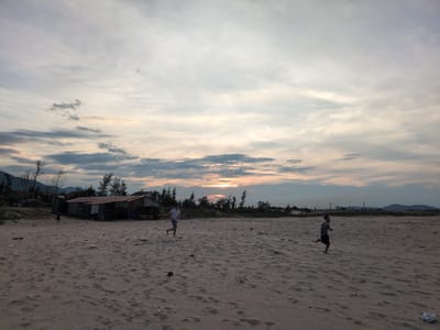A wide-angle, low-light photograph depicts a sandy beach in Hoành Sơn Ward, Vietnam, at dusk. The sky is dominated by dramatic clouds, with a sliver of the setting sun visible on the horizon, casting a warm glow through the atmospheric haze. In the mid-ground, a small, rustic building with a corrugated metal roof and open sides stands on the sand, flanked by sparse trees. A few people are present on the beach. To the left of the building, a dark-clothed figure is visible. Further out in the sandy expanse, a man in a white shirt and blue shorts is captured mid-stride, running to the left. To the right, a young boy in dark shorts and a patterned shirt is also running to the left. The beach itself is uneven and textured with sand ripples and scattered debris. In the background, a line of trees separates the beach from distant hills and what appear to be industrial structures. The overall impression is one of a serene, yet active, coastal scene at the close of day.