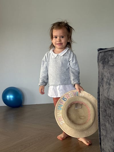 A full-body shot of a young child, approximately a toddler, standing indoors in Kharnavy, Russia. The child is looking towards the camera with a gentle smile, their light brown hair slightly disheveled. They are dressed in a white collared shirt under a grey knitted cardigan, with a partially visible reddish-brown clothing item on their lower body. Their bare feet are visible on the floor.
The child is holding a straw hat in their right hand, which has the word "SUMMER" embroidered in colorful letters (blue, pink, orange, green, light blue, pink) around its brim. The background consists of a plain light gray wall and a dark wooden floor with a herringbone pattern. A shiny blue balloon rests on the floor to the left of the child. A dark gray sofa or armchair is partially visible in the upper background. The natural lighting suggests the scene takes place during the daytime.