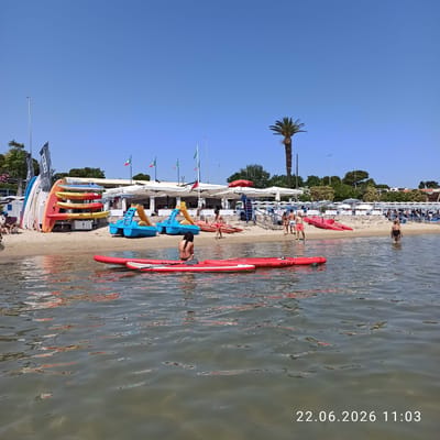 This media file captures a vibrant beach scene in Trani, Italy. The image is oriented vertically, with the sky occupying the top portion and the water and beach filling the rest. Several people are visible enjoying the beach and the water. Some are wading in the shallow water, while others are relaxing on the sandy shore. There are numerous red paddleboards and kayaks lined up on the beach and in the water, suggesting recreational activities are readily available. Beach umbrellas and chairs are set up along the shoreline, indicating a well-established beach resort.
Notable details include a palm tree on the left side of the frame, a characteristic element of Mediterranean landscapes. Italian flags are also visible on flagpoles, reinforcing the location. The water is clear and reflects the sunlight, creating a shimmering effect. The overall atmosphere appears to be lively and summery, suggesting a warm and sunny day. The time of day is indicated by the text overlay "22.06.2026 11:03", placing the scene in the late morning. The text on one of the paddleboards reads "AW-SURF 10'6 32"". The scene conveys a sense of leisure and outdoor enjoyment.