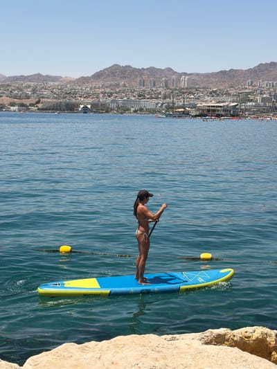 A woman is paddling a blue and yellow "WATTSUP" stand-up paddleboard in the waters of the Red Sea, near Eilat, Israel. The woman is wearing a dark cap and a patterned bikini. The paddleboard is tethered to a rocky shoreline. In the background, a city stretches along the coastline, with a backdrop of mountains under a clear blue sky. Yellow buoys are visible in the water.