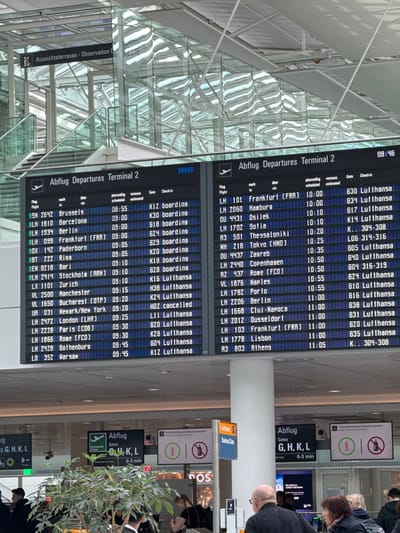 The image displays the interior of a modern airport terminal, identified as "Terminal 2 Abflug Departures," located at Munich Airport (Flughafen München Franz Josef Strauß) in Freising, Germany. Two large digital departure boards dominate the mid-ground, indicating the current time as 08:46 on the right board. These boards list various domestic and international flights, including destinations such as Brussels, Barcelona, Frankfurt, London, Paris, Hamburg, Tokyo, and Athens, with scheduled/estimated departure times ranging from 08:55 to 11:05, corresponding gates, and check-in statuses, with numerous flights identified as Lufthansa.
The terminal architecture features a high, vaulted ceiling constructed from a glass and metallic framework, allowing natural light into the space and creating reflections on the glass surfaces. Upper levels with glass railings and staircases are visible in the background, along with a sign pointing to "Aussichtsterrasse - Observation D." In the lower foreground, multiple individuals, predominantly adults, are present. A bald man in a dark jacket is centrally located in the lower right foreground, facing away from the camera. Other individuals are seen walking or standing, suggesting passenger movement. Green leafy plants in planters are positioned in the lower left and center foreground. Additional digital signs are visible, one indicating "Abflug Gates G, H, K, L 0-3 min" for Economy Class and another displaying "Business Class" with Lufthansa branding. Overhead signs feature iconography, including a crossed-out bottle, indicating restrictions. The scene depicts a functional and active airport environment during daylight hours.