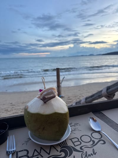 A whole, green coconut with its top cut off and a garnish of what appears to be a purple flower and a chocolate-covered item on a skewer is placed on a small white saucer. The coconut sits on a tray in the foreground, beside a fork to the left and a spoon to the right. The tray is on a table with a placemat featuring the logo "BAMBOO Club". In the background, a sandy beach stretches towards the ocean, with gentle waves lapping the shore. The sky above is cloudy with hints of pink and orange from a sunset. A wooden post stands in the sand near the water. The scene is set at the Bamboo Club in Ban Klong Haeng, Thailand.