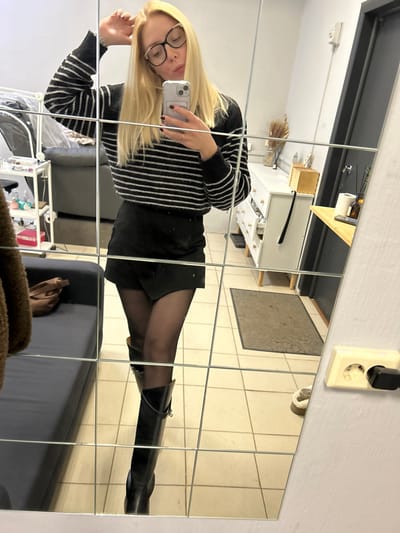 A blonde female is depicted in a full-body mirror selfie taken within an indoor residential or commercial space, specifically located in Voskresensk, Russia. The subject, appearing to be an adult, wears a black and white striped long-sleeved sweater, a black mini-skirt, dark sheer tights, and black knee-high boots with strap detailing. She holds a light-colored smartphone in her left hand, which is positioned centrally for the photograph, and her right hand is raised to touch the back of her head. Clear-framed eyeglasses are worn, and her lips are slightly pursed.

The reflection reveals a room with light-colored square floor tiles. To the subject's left, a dark gray sofa or armchair is partially visible, alongside a metal rolling cart containing various items. A brown, textured garment hangs to the far left. To the subject's right, a white dresser with multiple drawers and light wooden knobs is visible, adorned with a vase containing dried foliage, a wooden box, and a dark shoehorn. A wall-mounted wooden shelf holds a dark bottle and a paper roll. A dark grey doorway is also present. On the actual wall to the right of the mirror, a beige electrical outlet with a black plug inserted is visible. A dark grey rectangular mat lies on the floor in the background.