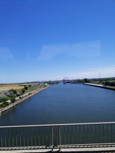 A wide-angle, eye-level shot captures a view of a large, calm body of water, likely a canal or river, under a clear, bright blue sky. In the foreground, a metal railing of a bridge or walkway is visible, casting shadows on the concrete below. The waterway stretches into the distance, bordered by concrete embankments and a path on the left side. Beyond the path, a dry, grassy field is seen, transitioning into green vegetation and scattered trees. In the far distance, a complex of industrial structures with tall cranes and buildings is apparent, suggesting a port or industrial area. The overall scene depicts a sunny day in an area that combines natural waterways with industrial development. The location is identified as Constanța, Romania.