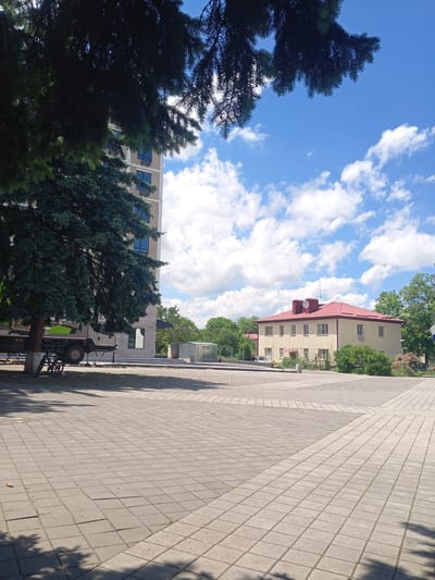 In the city of Yessentukskaya, Russia, a paved plaza is visible under a bright blue sky dotted with white clouds. To the left, a large evergreen tree casts shadows on the plaza. Behind the tree, a modern, light-colored multi-story building with blue-tinted windows stands tall. Next to the tree and partially obscured by it, a vehicle, possibly a truck with a green accent, is parked, and a bicycle leans against the tree. In the mid-ground, a yellow building with a red roof features multiple windows and what appears to be an air conditioning unit. Further in the background, lush green trees and bushes create a natural backdrop. A small, rectangular structure with glass walls, possibly a greenhouse, is also visible. The overall scene depicts a public outdoor space on a clear day, with elements of both modern and older architecture, along with natural greenery.