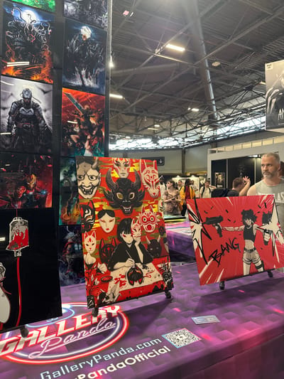 The image shows an indoor exhibition hall, likely at the Paris-Nord Villepinte Exhibition Centre in Villepinte, France, given the context of a convention or trade show. In the foreground, a table displays various artworks, primarily prints on easels. One prominent artwork features a collage of Japanese-style masks and two anime-style figures. To its right, another artwork depicts a woman holding two guns with the word "BANG" in bold lettering. The table itself is covered with a purple cloth featuring the "Gallery Panda" logo and a QR code.
In the background, more artworks are displayed on a vertical panel. These appear to be prints of fantasy or comic book characters, with one showing a figure resembling Batman and another featuring characters from Final Fantasy. Several people are visible in the mid-ground and background, browsing the stalls and interacting with the environment. The hall has a high ceiling with exposed industrial-style beams and lighting fixtures. The overall impression is of a vibrant convention setting focused on art and popular culture.