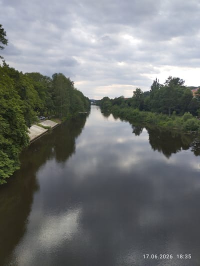 The image depicts a wide river flowing through a lush green landscape under a cloudy sky. On the left bank, dense trees overhang the water, with a concrete embankment and a small paved path visible between the trees and the water. Parked on the path are a white car and a parked motorcycle. The right bank is also lined with trees and greenery, with some buildings visible in the distance, suggesting an urban or semi-urban setting.

The river's surface is dark and reflects the overcast sky and the surrounding trees, creating a mirror-like effect. The clouds are thick and grey, indicating a potentially damp or overcast day. The light suggests it is late afternoon or early evening, with the timestamp "17.06.2026 18:35" confirming this. The overall mood is calm and serene, with the natural landscape dominating the scene. There is no discernible activity taking place, apart from the implied presence of the parked vehicles on the left bank. The location is identified as Karlovy Vary, Czechia, which aligns with the picturesque river and green surroundings.