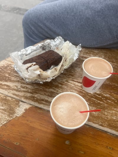 The image shows a top-down view of a wooden table with two cups of hot chocolate and a chocolate pastry. The pastry is wrapped in foil and paper, and appears to be a dense, dark chocolate cake or brownie. One cup of hot chocolate is closer to the foreground, and the other is slightly further back. Both cups are white with red accents and have red straws. In the background, a person's legs are visible, covered in grey sweatpants. The table appears to be outdoors, as indicated by the lighting and the weathered appearance of the wood. The location is identified as Maracay, Venezuela.