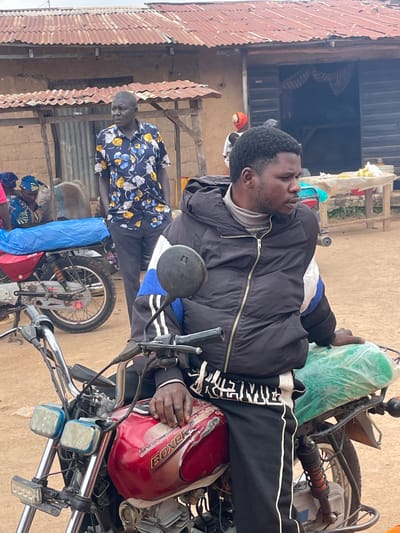 In the city of Wang, Nigeria, a man with dark skin and a beard sits on a red motorcycle. He is wearing a black puffer jacket with a gray turtleneck underneath, and black pants with white stripes. His right hand rests on the motorcycle's seat. Behind him, another motorcycle with a blue tarp covering its seat is parked. In the background, several people are visible. A man in a floral shirt stands near a building with a corrugated metal roof. To the left, two women wearing headscarves are seen. A wooden table with various items is positioned in front of a dark doorway. The ground is dusty and appears to be dirt.