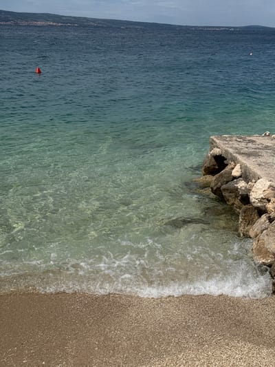 The image shows a view of the Adriatic Sea from the shoreline in Dugi Rat, Croatia. The location is pinpointed by the coordinates 43.446 latitude and 16.634 longitude. The image captures a section of the beach with a concrete pier jutting out into the water. The water is clear and turquoise, with a sandy bottom visible in some areas. Small white waves are lapping onto the sandy beach. There is a small, red buoy visible in the distance. The sky is a hazy blue with a few clouds. The image appears to have been taken during the day in bright sunlight.