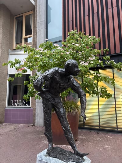 This forensic image captures a street scene in Arnhem, Netherlands, specifically near Koningstraat 24-26, featuring the sculpture "De Klopper" by Jan Wolkers.
The central subject is a dark, textured bronze statue of a male figure, standing approximately human height. The figure is depicted in a stooped, somewhat contorted pose, with its left hand resting on its hip and its right arm extended downwards, fingers slightly curled. The statue rests on a polished light grey marble base, which is set upon reddish-brown brick paving stones.
Behind the statue, a medium-sized tree with vibrant green leaves is in bloom, displaying numerous light pink to white blossoms. The tree partially obscures the background structures.
To the left of the statue, a multi-story building facade is visible. It features light brown brickwork and a prominent vertical concrete structural element. A window with a light-colored wooden frame and interior curtains is present on an upper floor. Below this, a commercial storefront has a large white-framed window. Prominently displayed in white capital letters on the window is "ULLA POPKEN." Above this, partially obscured by tree foliage, faint lettering reads "RM Huurhuizen" and the local phone area code "026" is discernable. Below the shop window, the building features a section of purple tiled facade.
To the right of the statue, a modern building showcases a facade composed of alternating dark and reddish-brown vertical panels. Below this, a large window reflects strong horizontal bands of yellow and orange light, suggesting internal lighting or blinds. A large, cylindrical planter with a rusted reddish-brown finish containing green grassy foliage is positioned behind the statue's legs, to the left of the reflective window.
The ground surface throughout the visible area consists of uniformly laid reddish-brown rectangular brick pavers, showing some minor wear and scattered small weeds. The overall lighting suggests an overcast or late afternoon condition.