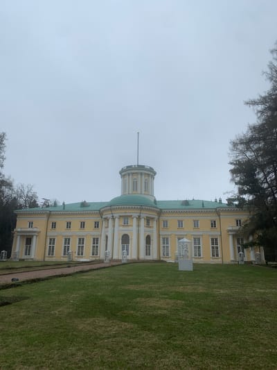 The image shows the main building of the Arkhangelskoye Estate in Russia. The building is predominantly yellow with white architectural detailing and a green roof. It features a central rotunda with a spire. The façade is lined with numerous windows, and there are decorative elements such as columns and statues. The building is surrounded by a large expanse of green lawn. Trees are visible on the left and in the foreground. The sky is overcast and grey. No people are clearly visible.
