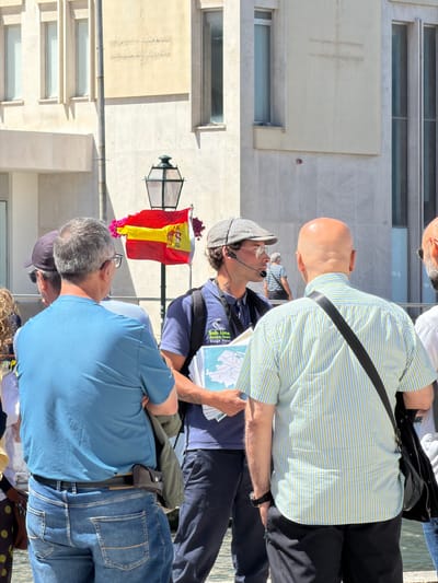 In Aveiro, Portugal, on a sunny day, a tour guide wearing a flat cap and headset is addressing a group of tourists. The guide holds a map, indicating they are leading a tour. The group is standing on a paved area, possibly a street or promenade, with a building in the background. A Spanish flag is visible, attached to a lamppost, along with decorative pink flowers. The tourists are casually dressed, suggesting a typical sightseeing activity.
