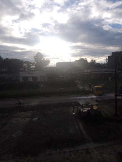 The scene depicts an outdoor urban or semi-urban environment in Njoro ward, Kenya, characterized by challenging lighting conditions due to a heavily overexposed sky and a dark foreground. A bright light source, consistent with the sun, is positioned high in the upper-center, illuminating scattered clouds.
In the background, silhouetted buildings, some appearing to be under construction with visible scaffolding, extend across the horizon. Multiple power lines traverse the upper and mid-sections. A distinct white building with dark windows and roof is visible in the mid-left background, partially obscured by trees.
The midground features an unpaved or partially paved road running horizontally. One individual on a bicycle, wearing a bright orange or red top, travels from left to right on this road. Further right on the road, a yellow vehicle, possibly a small bus or truck, is stationary or moving slowly, positioned next to a reflective patch on the ground, possibly water.
In the right mid-to-foreground, a small, covered stall or cart structure with a light-colored roof is situated off the road. At least two individuals are visible near this structure, one standing adjacent to it and another slightly behind. The foreground consists of dark, uneven ground, characterized by dirt or gravel, with several small piles of material. Utility poles are present throughout the midground and background, supporting the power lines.
