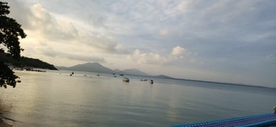 A wide-angle, eye-level shot captures a serene bay at Ao Wiang Wan, Thailand. The calm, greyish-blue water stretches towards a hazy horizon, punctuated by several small boats anchored or slowly moving. In the distance, lush green, mountainous islands rise, their peaks softened by a light mist. The sky above is a canvas of muted grey and white clouds, with hints of pale yellow and orange suggesting either early morning or late afternoon light. The left side of the frame features a dark, silhouetted tree branch extending into the sky, and a wooden pier with several structures and figures. A portion of a blue and white patterned walkway or floating dock is visible in the lower right corner, with a person partially visible on it. The overall scene is tranquil and evokes a sense of peacefulness.