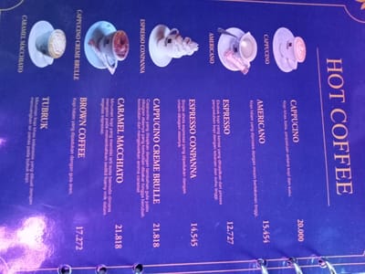 The image displays a tilted, dark blue menu for "HOT COFFEE," likely from a cafe or restaurant. The menu is bound with metal rings visible at the bottom. Small, illustrative images of various coffee drinks, served in cups and saucers, are placed next to their respective names.

The menu lists several coffee options with descriptions and prices in Indonesian Rupiah (indicated by the ".000" and decimal format). From right to left, the items include:

*   **CAPPUCINO** (20.000): Described as a classic Italian coffee, a blend of coffee and milk.
*   **AMERICANO** (15.454): Black coffee processed with a high-pressure machine.
*   **ESPRESSO** (12.727): A concentrated coffee extract made through high-pressure and temperature brewing.
*   **ESPRESSO CONPANNA** (14.545): Single-shot espresso topped with cream.
*   **CAPPUCINO CREME BRULLE** (21.818): Cappuccino served with palm sugar on top, torched until caramelized to release a caramel aroma.
*   **CARAMEL MACCHIATO** (21.818): A coffee drink described as "stained milk," referring to the addition of a little foamy milk to a glass of espresso.
*   **BROWN COFFEE** (17.272): Black coffee mixed with palm sugar.
*   **TUBRUK** (17.272): A traditional Indonesian coffee made by pouring hot water directly onto coffee grounds.

The overall color scheme is dark blue with white and gold text, contributing to a sleek appearance. The lighting appears artificial, suggesting an indoor setting.
