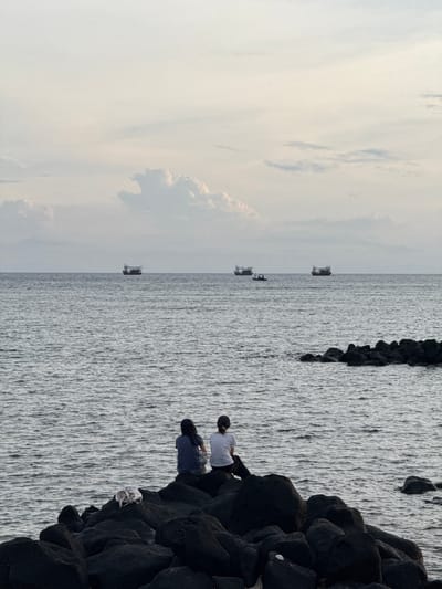 Two individuals are seated on dark volcanic rocks, facing the ocean. The person on the left wears a dark blue t-shirt and dark shorts. The person on the right wears a white t-shirt and dark pants, with a dark cap. Between them on the rocks is a white plastic bag. The ocean is calm with gentle ripples, and several fishing boats are visible in the distance on the horizon under a cloudy sky. This scene appears to be from the coast of Lý Sơn Island, Vietnam.