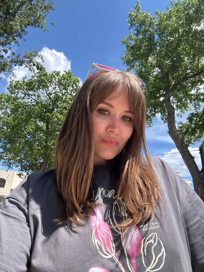 A woman with shoulder-length brown hair is taking a selfie outdoors. She has fair skin, light eyes, and is wearing a gray t-shirt with a floral design. Part of pink sunglasses is visible above her forehead. The background consists of green trees, a blue sky with white clouds, and a building partially visible on the right. The photo does not provide enough information to determine a specific location in Yevpatoriya, Ukraine.
